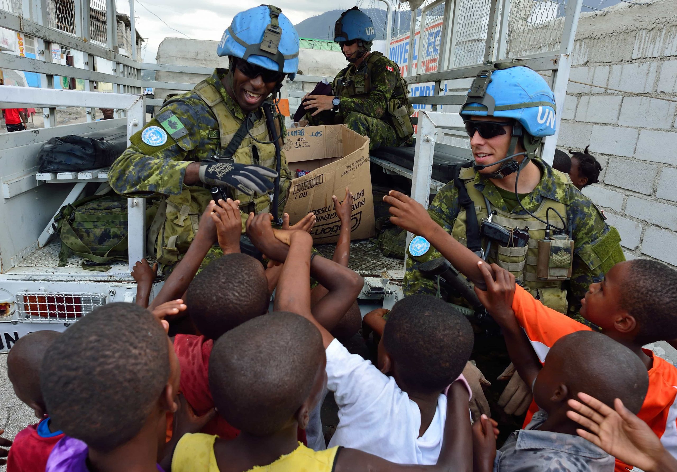 A crowd of Black children reach out to a smiling Black man in a peacekeeping uniform, while a white man in a peacekeeping uniform watches.